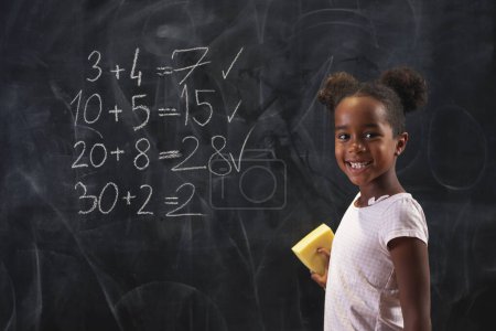 Photo for Beautiful mixed race schoolgirl standing in front of a chalkboard, solving math equations, practicing summation - Royalty Free Image