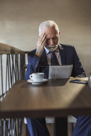 Photo for Senior businessman sitting at a restaurant table, drinking coffee and reading business news using a tablet computer, pensive and serious - Royalty Free Image