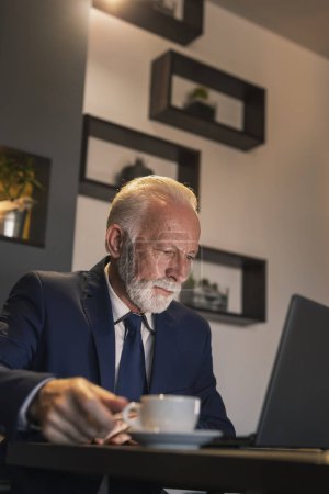 Téléchargez les photos : Homme d'affaires senior dans un restaurant, travaillant sur un ordinateur portable et prenant une tasse de café - en image libre de droit