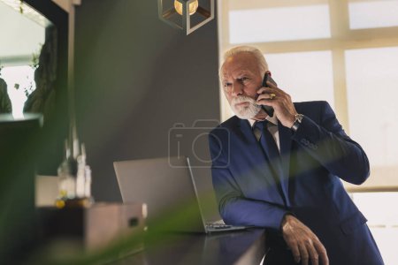 Téléchargez les photos : Homme d'affaires senior travaillant dans un bureau moderne, ayant une conversation téléphonique tout en travaillant sur un ordinateur portable - en image libre de droit