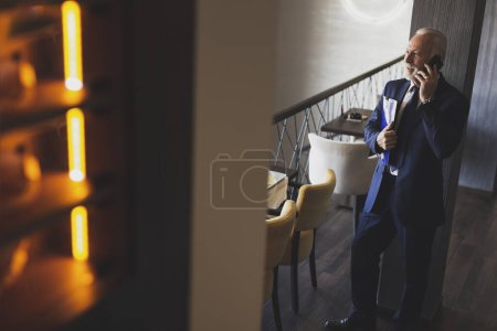 Téléchargez les photos : Homme d'affaires principal debout dans un restaurant moderne d'immeuble de bureaux, ayant une conversation téléphonique et tenant quelques dossiers de document - en image libre de droit