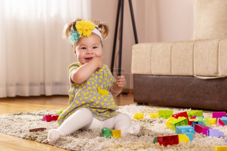 Photo for Beautiful little baby girl, sitting on a carpet on the nursery floor, chewing her hands and smiling - Royalty Free Image