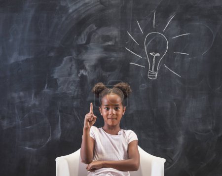 Photo for Elementary school girl sitting in chair in front of a chalkboard in classroom, happy after coming up with an idea, holding index finger up with light bulb drawn on the blackboard behind her - Royalty Free Image