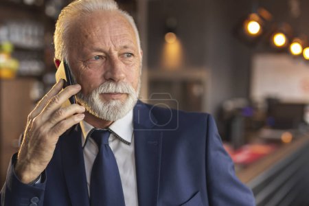 Téléchargez les photos : Homme d'affaires principal debout à côté d'un comptoir moderne de restaurant d'immeuble de bureaux, parlant au téléphone - en image libre de droit