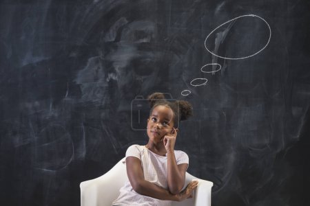 Foto de Hermosa chica de la escuela primaria de raza mixta sentada en silla frente a una pizarra pensativa y seria, pensando, con nubes dibujadas sobre su cabeza - Imagen libre de derechos