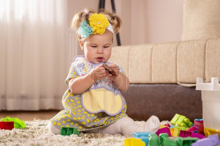 Foto de Hermosa niña, sentada en una alfombra en el piso del vivero, jugando y masticando bloques de juguetes - Imagen libre de derechos