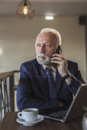 Téléchargez les photos : Homme d'affaires senior travaillant dans un bureau moderne, ayant une conversation téléphonique tout en travaillant sur un ordinateur portable - en image libre de droit