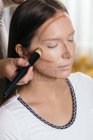 Foto de Maquillaje artista aplicando base de polvo de cara líquida a la cara de un cliente femenino y mezclando contornos - Imagen libre de derechos