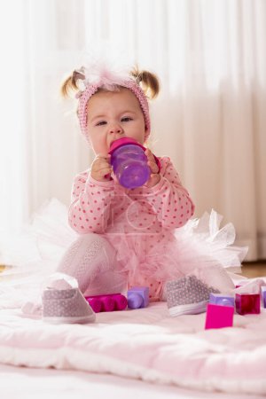 Foto de Hermosa niña pequeña con falda rosa tutú, sentada en el piso del vivero y bebiendo agua de un biberón - Imagen libre de derechos