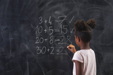 Téléchargez les photos : Belle écolière métisse debout devant un tableau, résolvant des équations mathématiques, pratiquant la sommation - en image libre de droit