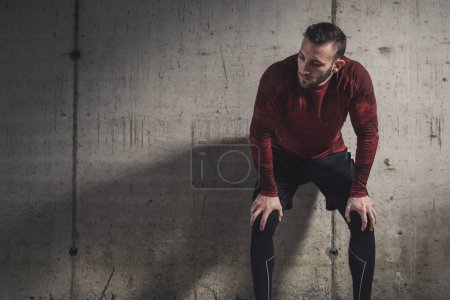 Foto de Hombre atlético musculoso usando ropa deportiva, tomando un descanso de entrenamiento, de pie y apoyado en una pared de hormigón, pensativo - Imagen libre de derechos