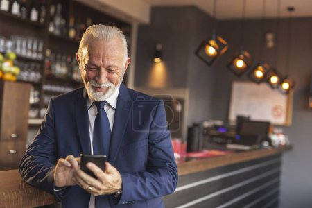 Téléchargez les photos : Homme d'affaires senior debout à côté d'un comptoir moderne de restaurants de bureaux, tapant un message texte à l'aide d'un téléphone intelligent - en image libre de droit