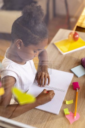 Téléchargez les photos : Belle petite fille qui s'amuse à placer des notes collantes colorées sur une fenêtre ouverte et à écrire et dessiner dans un carnet - en image libre de droit