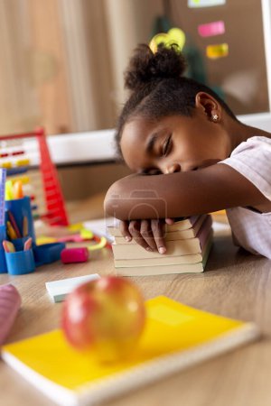 Téléchargez les photos : Belle petite fille fatiguée de faire ses devoirs, dormant à son bureau, utilisant une pile de livres comme oreiller - en image libre de droit