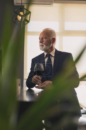 Photo for Senior businessman standing next to a counter in a restaurant, drinking wine while on a lunch break - Royalty Free Image
