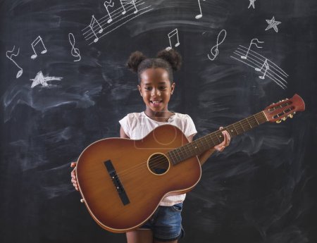 Photo for Beautiful mixed race elementary school girl standing in front of a chalkboard in classroom, playing the guitar and singing while in music class, with musical note symbols drawn on the blackboard - Royalty Free Image