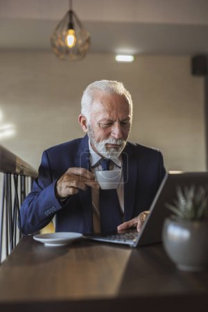 Téléchargez les photos : Homme d'affaires senior assis à une table de restaurant, prendre une tasse de café et lire des nouvelles sur ordinateur portable - en image libre de droit