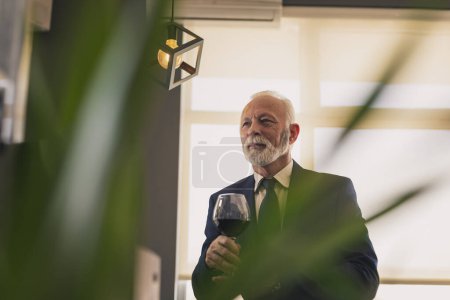 Photo for Senior businessman standing next to a counter in a restaurant, drinking wine while on a lunch break - Royalty Free Image