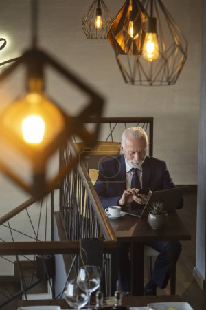 Photo for High angle view of senior businessman sitting in modern office building restaurant, using a smart phone and working on laptop computer - Royalty Free Image