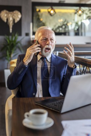 Téléchargez les photos : Homme d'affaires principal travaillant dans un bureau moderne, ayant une conversation téléphonique tout en travaillant sur un ordinateur portable, en colère et mécontent - en image libre de droit