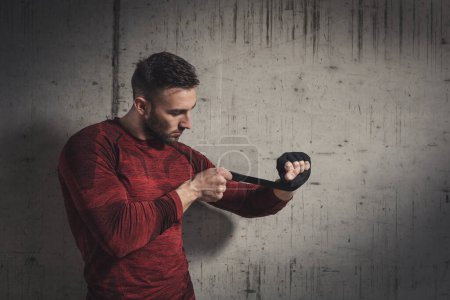 Téléchargez les photos : Homme musclé enveloppant des bandages sur les mains avant un entraînement de boxe ; boxeur se préparant pour un combat - en image libre de droit