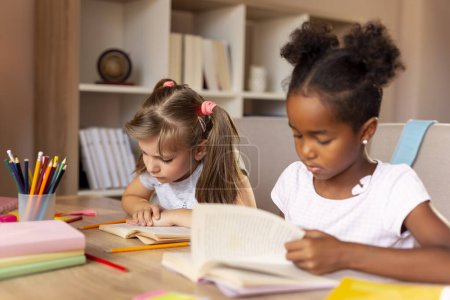 Téléchargez les photos : Deux petites filles assises à un bureau à la maison, lisant des livres et étudiant pour l'école. Concentrez-vous sur la fille à gauche - en image libre de droit
