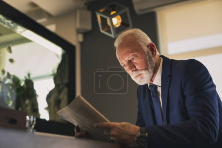 Téléchargez les photos : Homme d'affaires senior debout à côté d'un comptoir dans un café, lisant un journal - en image libre de droit