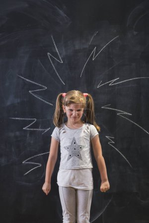 Photo for Angry schoolgirl standing in front of drawn high voltage signs on blackboard behind her with furious face expression and clenched fists - Royalty Free Image