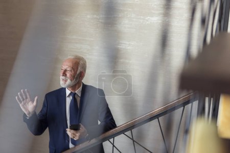 Téléchargez les photos : Un homme d'affaires chevronné monte les escaliers d'un immeuble de bureaux moderne, se rend sur son lieu de travail, parle à un collègue et utilise un téléphone intelligent - en image libre de droit
