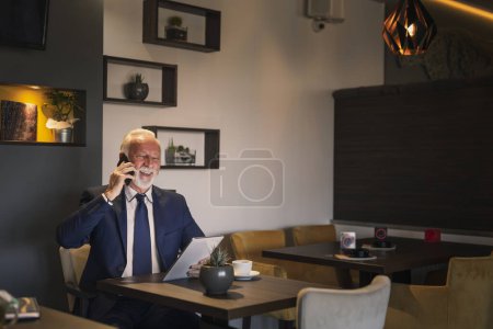 Photo for Senior businessman in a restaurant, having a phone conversation while working on a tablet computer - Royalty Free Image