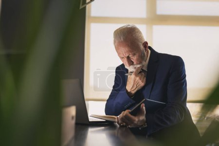 Foto de Hombre de negocios sénior en una oficina moderna trabajando en una computadora portátil y leyendo notas de su planificador, pensativo y serio - Imagen libre de derechos