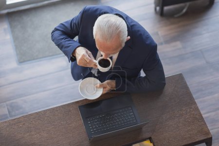 Téléchargez les photos : Portrait en grand angle d'un homme d'affaires âgé debout près d'un comptoir de restaurant, buvant du café et travaillant sur un ordinateur portable - en image libre de droit