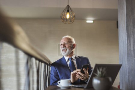 Téléchargez les photos : Homme d'affaires senior assis à une table de restaurant, prenant une tasse de café, travaillant sur un ordinateur portable et tapant un message texte à l'aide d'un téléphone intelligent - en image libre de droit