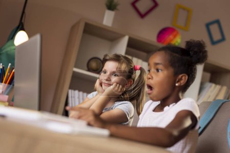 Foto de Dos hermosas niñas sentadas en un escritorio, viendo dibujos animados en una computadora portátil y divirtiéndose - Imagen libre de derechos
