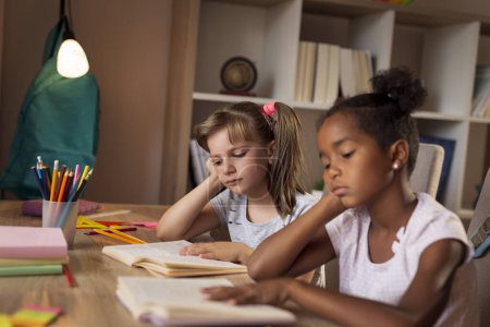 Photo for Two little girls sitting at a desk at home, reading books and studying for an exam, tired and unhappy. Focus on the girl on the left - Royalty Free Image