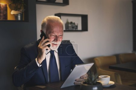 Téléchargez les photos : Homme d'affaires principal dans un restaurant, ayant une conversation téléphonique tout en travaillant sur un ordinateur portable - en image libre de droit