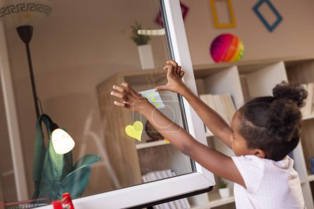 Photo for Beautiful little girl having fun placing colorful sticky notes on an opened window and writing and drawing on them - Royalty Free Image