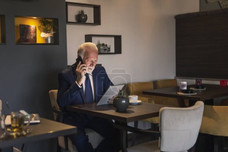 Photo for Senior businessman sitting at a restaurant table, having a phone conversation while working on a tablet computer - Royalty Free Image