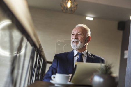 Téléchargez les photos : Homme d'affaires principal assis dans un restaurant moderne d'immeuble de bureaux, buvant du café et signant la documentation, satisfait et heureux - en image libre de droit