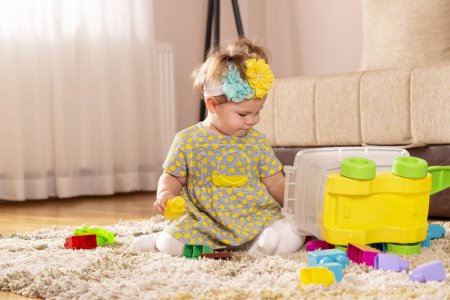 Photo for Beautiful little baby girl, sitting on a carpet on the nursery floor, playing with toy blocks, pensive and serious - Royalty Free Image