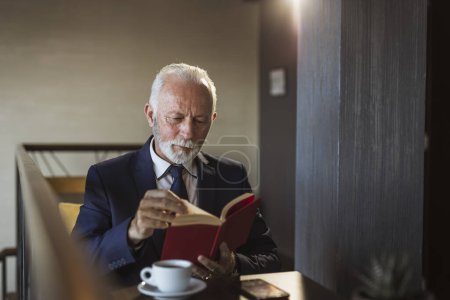Téléchargez les photos : Homme d'affaires senior assis à une table de restaurant, lisant un livre et buvant du café - en image libre de droit
