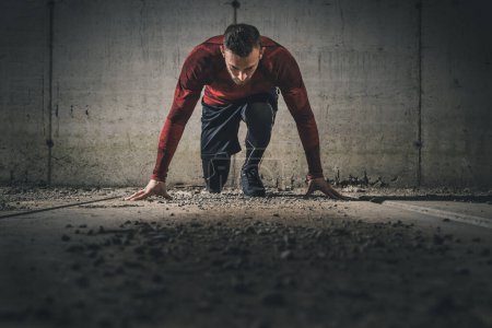 Photo for Young, athletic man working out on a construction site in front of a concrete wall, getting ready for sprinting - Royalty Free Image
