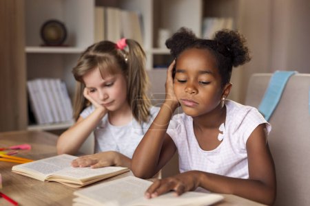 Foto de Dos niñas sentadas en un escritorio en casa, leyendo libros y estudiando para un examen, cansadas e infelices - Imagen libre de derechos