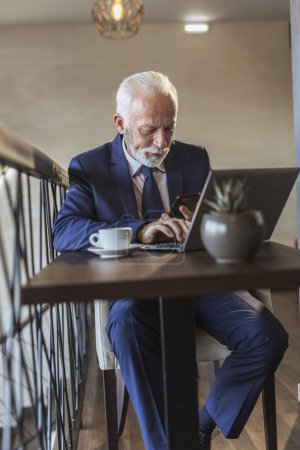 Photo for Senior businessman sitting at a restaurant table, having a cup of coffee, working on a laptop computer and typing a text message using smart phone - Royalty Free Image