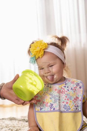 Foto de Hermosa niña siendo alimentada por su madre, comiendo papanatas vegetales y sonriendo, untada por toda la cara, mirando divertido en un tazón vacío - Imagen libre de derechos