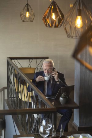 Téléchargez les photos : Vue en angle élevé d'un homme d'affaires senior dans un restaurant moderne d'immeuble de bureaux, ayant une conversation téléphonique tout en travaillant sur un ordinateur portable - en image libre de droit