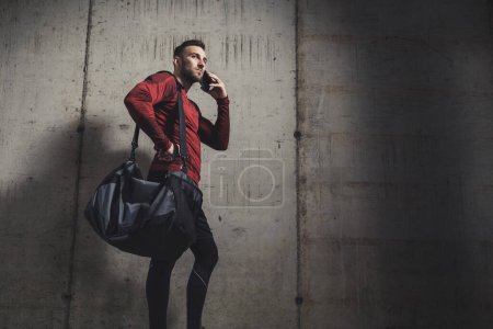 Foto de Hombre atlético musculoso llevando una bolsa de gimnasio y usando ropa deportiva, teniendo una conversación telefónica después de un duro entrenamiento - Imagen libre de derechos
