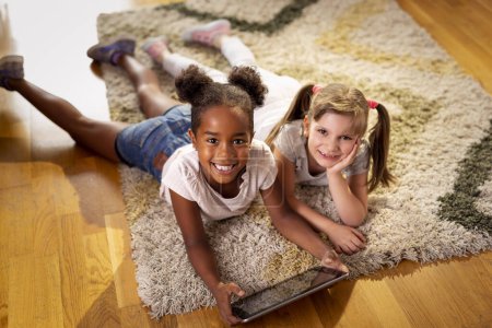 Foto de Vista de ángulo alto de dos niñas que yacen en el piso de la sala de juegos, jugando juegos en una tableta y divirtiéndose. Concéntrate en la chica de la izquierda - Imagen libre de derechos