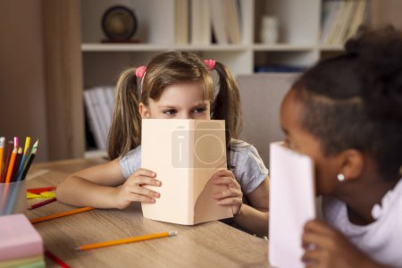Photo for Two little girls sitting at a desk at home, hiding behind books and peeking; first graders doing their homework and reading books - Royalty Free Image