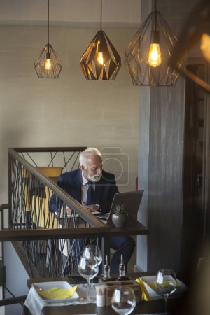 Téléchargez les photos : Vue en angle élevé d'un homme d'affaires âgé assis à une table de restaurant, travaillant sur un ordinateur portable, écrivant dans un planificateur et buvant du café - en image libre de droit